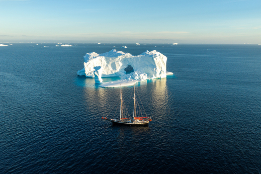 Aerial view of a large arch iceberg floating in the deep blue ocean with a classic sailing boat navigating nearby in Greenland. Aerial view of a large arch iceberg floating in the deep blue ocean with a classic sailing boat navigating nearby in Greenland.