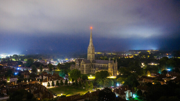 Aerial view of the illuminated Salisbury Cathedral spire rising above the historic city rooftops and glowing street lights at night in Salisbury England United Kingdom. Aerial view of the illuminated Salisbury Cathedral spire rising above the historic city rooftops and glowing street lights at night in Salisbury England United Kingdom.