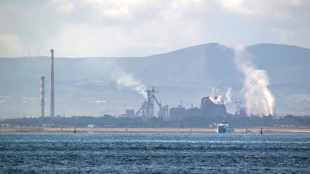 View of an industrial complex with tall smokestacks emitting smoke and a ferry crossing the water with mountains in the background Barreiro Setubal Portugal.