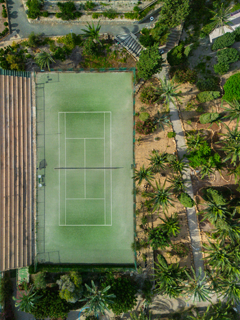 Aerial view of a green tennis court surrounded by lush palm trees and landscaped gardens in Spain.
