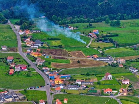 Aerial view of a rural village with colorful houses green agricultural fields and smoke rising from a fire near Cudillero Asturias Spain.
