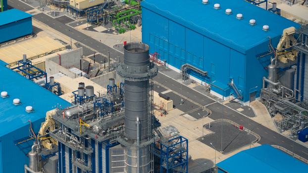 Aerial view of the Gulf PD Power Plant featuring large blue industrial buildings a tall exhaust stack and intricate piping systems under bright daylight in Map Yang Phon Rayong Thailand.