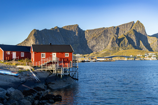 View of the iconic red rorbuer cabins on stilts along the rocky coastline with dramatic mountains rising in the background and a bridge spanning the water in Hamnoy Nordland Norway.
