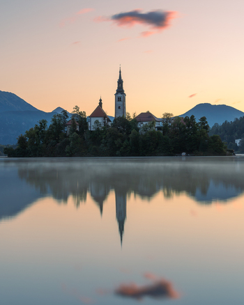 View of the Pilgrimage Church of the Assumption of Maria on Lake Bled with its reflection in the calm water at sunset surrounded by mountains in Bled Radovljica Slovenia.