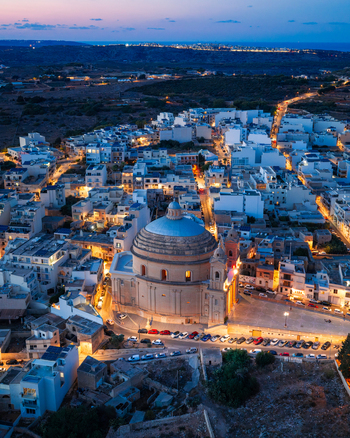 Aerial view of the Parish Church of the Assumption of the Blessed Virgin Mary into Heaven illuminated at twilight surrounded by the dense urban landscape of Mgarr Northern Region Malta.