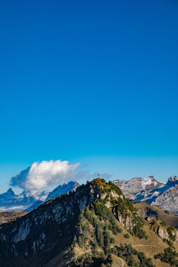 View of the Swiss Alps peak with a summit cross rocky slopes scattered trees and distant snow capped mountains under a clear blue sky in Airolo Ticino Switzerland. View of the Swiss Alps peak with a summit cross rocky slopes scattered trees and distant snow capped mountains under a clear blue sky in Airolo Ticino Switzerland.