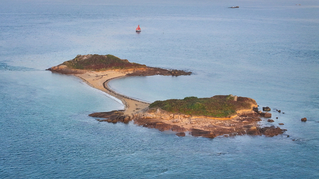Aerial view of a thin land bridge connecting two rocky verdant islands amidst the vast shimmering sea a solitary sailboat drifting in the distance Carantec Brittany France. Aerial view of a thin land bridge connecting two rocky verdant islands amidst the vast shimmering sea a solitary sailboat drifting in the distance Carantec Brittany France.