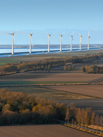 Aerial view of a row of modern wind turbines standing along a dike next to agricultural fields and a calm sea under a clear blue sky Netherlands.