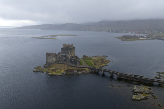 Aerial view of Eilean Donan Castle on its tidal island with the stone bridge and misty mountains under a cloudy sky Dornie Scotland United Kingdom.