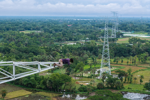 Trishal Upazila Bangladesh 13 May 2026 Aerial view of a man lying on the arm of a high voltage electricity pylon overlooking a lush green landscape with fields and trees. Trishal Upazila Bangladesh 13 May 2026 Aerial view of a man lying on the arm of a high voltage electricity pylon overlooking a lush green landscape with fields and trees.
