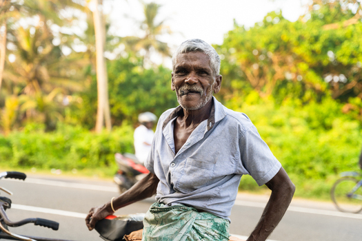 Sri Lanka 21 January 2026 View of an elderly man smiling while standing with his bicycle on a roadside surrounded by lush tropical greenery.