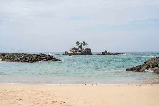 View of a small island temple surrounded by turquoise ocean water and stone breakwaters from a sandy beach in Dodanduwa Southern Province Sri Lanka. View of a small island temple surrounded by turquoise ocean water and stone breakwaters from a sandy beach in Dodanduwa Southern Province Sri Lanka.