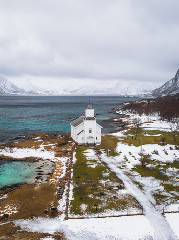 Aerial view of the white Gimsøy Church standing proudly against the backdrop of the turquoise sea and snow dusted mountains Gimsøysand Nordland Norway. Aerial view of the white Gimsøy Church standing proudly against the backdrop of the turquoise sea and snow dusted mountains Gimsøysand Nordland Norway.