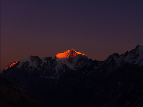 Aerial view of a snow capped Himalayan mountain peak illuminated by the warm golden light of sunrise against a dark twilight sky in Nepal.