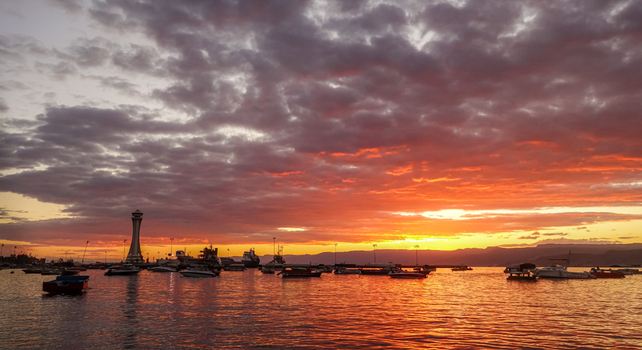 View of a harbor with numerous small boats and a tall tower silhouetted against a vibrant orange and purple sunset sky over the calm water in Jordan. View of a harbor with numerous small boats and a tall tower silhouetted against a vibrant orange and purple sunset sky over the calm water in Jordan.