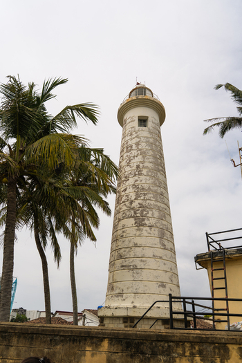 View of the historic Galle Lighthouse standing tall beside palm trees under a cloudy sky in Galle Southern Province Sri Lanka.