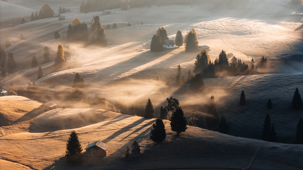 Aerial view of Alpe di Siusi with wooden cabins and pine trees scattered across rolling hills covered in morning mist under golden sunlight in Castelrotto Trentino Alto Adige Italy. Aerial view of Alpe di Siusi with wooden cabins and pine trees scattered across rolling hills covered in morning mist under golden sunlight in Castelrotto Trentino Alto Adige Italy.