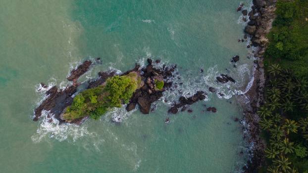 Aerial view of the rocky outcrop of Pantai Karang Agung with lush green trees surrounded by turquoise sea waves and a palm fringed coastline in Ayah Central Java Indonesia. Aerial view of the rocky outcrop of Pantai Karang Agung with lush green trees surrounded by turquoise sea waves and a palm fringed coastline in Ayah Central Java Indonesia.