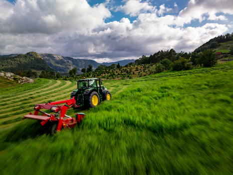 Aerial view of a green tractor with a red mower attachment harvesting grass for silage production in a lush green field with mountains under a cloudy sky in Rogaland Norway.