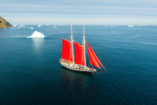 Aerial view of a classic sailboat with vibrant red sails navigating through deep blue Arctic waters surrounded by scattered icebergs in Greenland. Aerial view of a classic sailboat with vibrant red sails navigating through deep blue Arctic waters surrounded by scattered icebergs in Greenland.