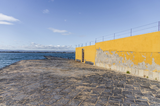 View of a weathered yellow wall meets the textured stone ground near the tranquil blue water a peaceful scene bathed in sunlight Ortigia Sicily Italy.