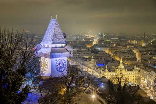 View of the clock tower standing majestically against the cityscape its face illuminated contrasting with the dark sky and snow touched trees Graz Styria Austria. View of the clock tower standing majestically against the cityscape its face illuminated contrasting with the dark sky and snow touched trees Graz Styria Austria.
