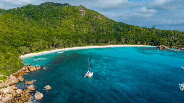 Aerial view of Anse Reunion beach with a catamaran anchored in turquoise water surrounded by lush green hills and granite rocks in Anse Reunion La Digue Seychelles.
