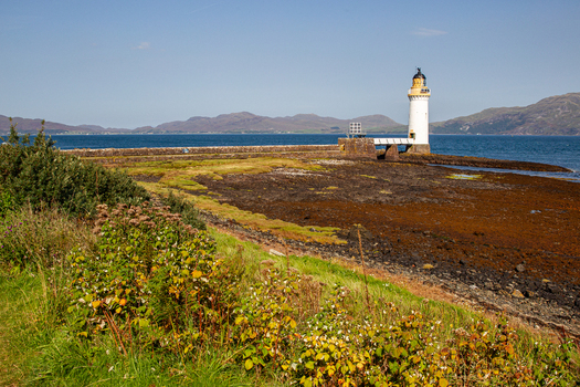 View of Tobermory Lighthouse standing on the rocky coastline surrounded by green vegetation and the blue waters of the Sound of Mull with distant hills Isle of Mull Scotland United Kingdom. View of Tobermory Lighthouse standing on the rocky coastline surrounded by green vegetation and the blue waters of the Sound of Mull with distant hills Isle of Mull Scotland United Kingdom.