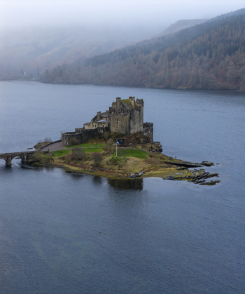 Aerial view of Eilean Donan Castle on its tidal island surrounded by calm waters and misty forested hills in Dornie Scotland United Kingdom.