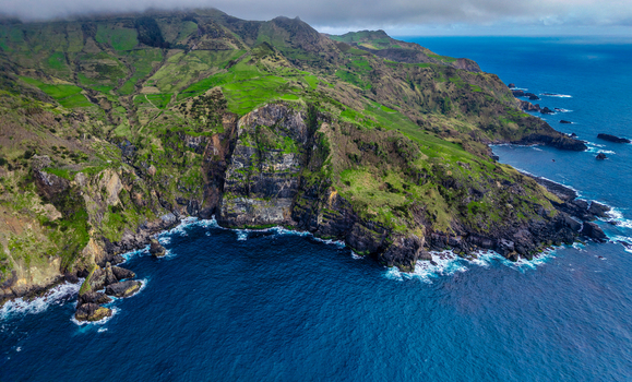 Aerial view of rugged cliffs rise dramatically from the deep blue sea fringed by white crested waves and verdant green slopes Faj&atilde; Grande Azores Portugal.