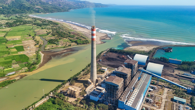 Aerial view of a power plant with a tall chimney situated near the coastline surrounded by lush green rice fields and the blue sea in Indonesia.