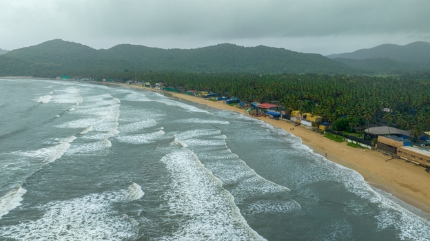 Aerial view of Palolem Beach with gentle waves rolling onto the sandy shore framed by lush palm trees and distant green hills in Canacona Goa India.