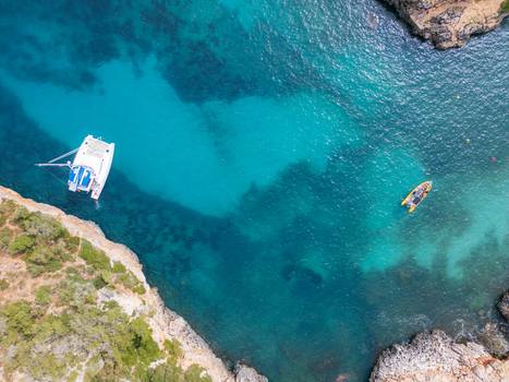Aerial view of Cala Sa Nau with a white catamaran and a yellow boat anchored in turquoise waters surrounded by rocky cliffs Felanitx Balearic Islands Spain. Aerial view of Cala Sa Nau with a white catamaran and a yellow boat anchored in turquoise waters surrounded by rocky cliffs Felanitx Balearic Islands Spain.