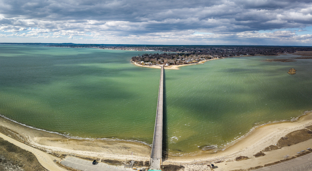 Aerial view of the Powder Point Bridge stretching across the turquoise waters to a wooded peninsula under a sky of textured clouds Duxbury Massachusetts United States.