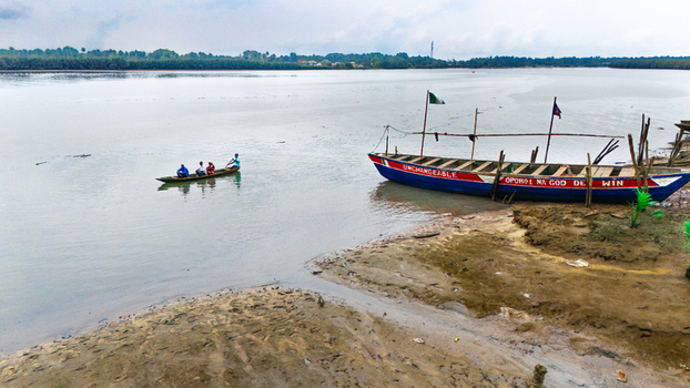 Aerial view of a wooden boat moored on the muddy riverbank with a small canoe carrying people paddling across the calm water in Minima Rivers Nigeria.
