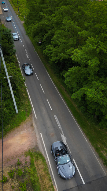 Aerial view of luxury sports cars driving along a paved road bordered by dense green trees and foliage in La Chapelle de Guinchay Bourgogne Franche Comte France.