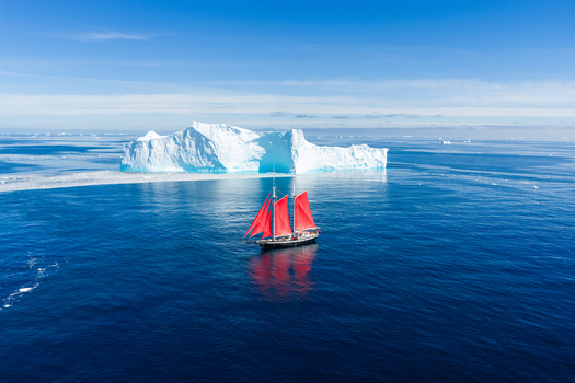 Aerial view of a sailboat with vibrant red sails navigating the deep blue ocean near a massive white iceberg in Greenland. Aerial view of a sailboat with vibrant red sails navigating the deep blue ocean near a massive white iceberg in Greenland.