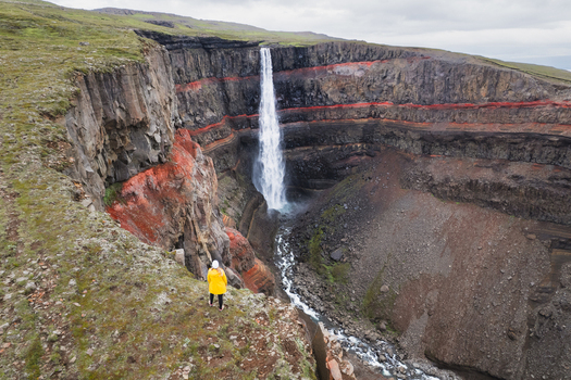 Aerial view of Hengifoss waterfall featuring red clay layers and a person in a yellow jacket on the cliff edge in Egilsstadir Fljotsdalshreppur Iceland. Aerial view of Hengifoss waterfall featuring red clay layers and a person in a yellow jacket on the cliff edge in Egilsstadir Fljotsdalshreppur Iceland.