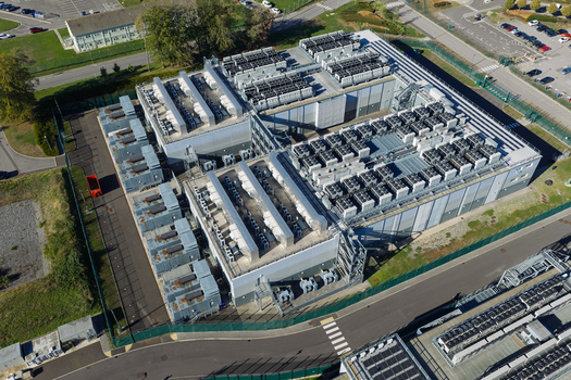 Aerial view of a large data center facility with rows of cooling units and industrial equipment on the roof surrounded by security fencing and parking areas in the United Kingdom.