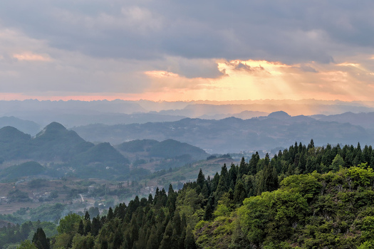 Aerial view of rolling hills and dense evergreen forests under a dramatic sky with sunbeams breaking through the clouds over Gaoxing Village Guizhou China.