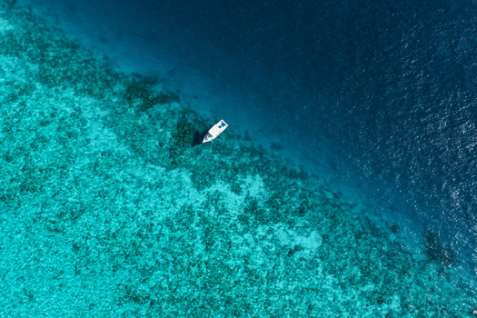 Aerial view of a small white boat floating in clear turquoise water above a vibrant coral reef with a sharp transition to the deep blue ocean in Vashafaru Haa Alif Atoll Maldives. Aerial view of a small white boat floating in clear turquoise water above a vibrant coral reef with a sharp transition to the deep blue ocean in Vashafaru Haa Alif Atoll Maldives.