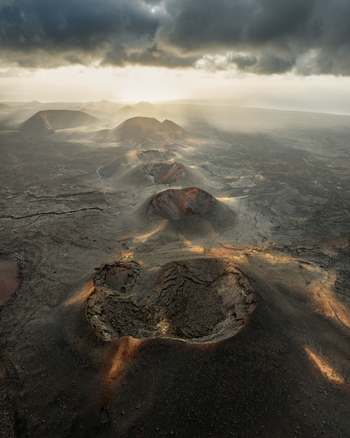 Aerial view of the volcanic craters of Timanfaya National Park and vast lava fields under a dramatic sky with sunbeams in Lanzarote Canarias Spain. Aerial view of the volcanic craters of Timanfaya National Park and vast lava fields under a dramatic sky with sunbeams in Lanzarote Canarias Spain.