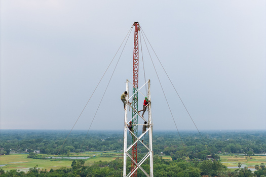 Trishal Upazila Bangladesh 13 May 2026 Aerial view of workers climbing a tall metal tower structure under construction above a lush green rural landscape. Trishal Upazila Bangladesh 13 May 2026 Aerial view of workers climbing a tall metal tower structure under construction above a lush green rural landscape.
