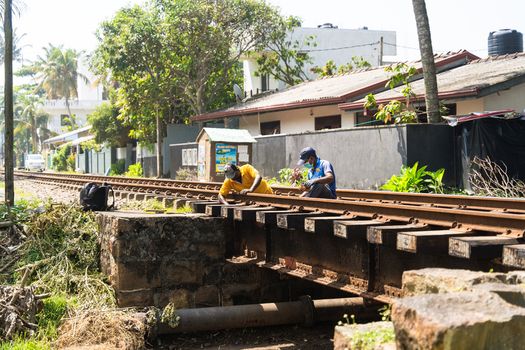 Sri Lanka 31 January 2026 View of two railway workers performing maintenance on a track bridge near residential houses and palm trees.