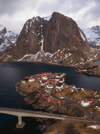 Aerial view of red roofed cabins clustered on a rocky islet beneath the dramatic silhouette of a snow dusted mountain Sakris&oslash;ya Nordland Norway.
