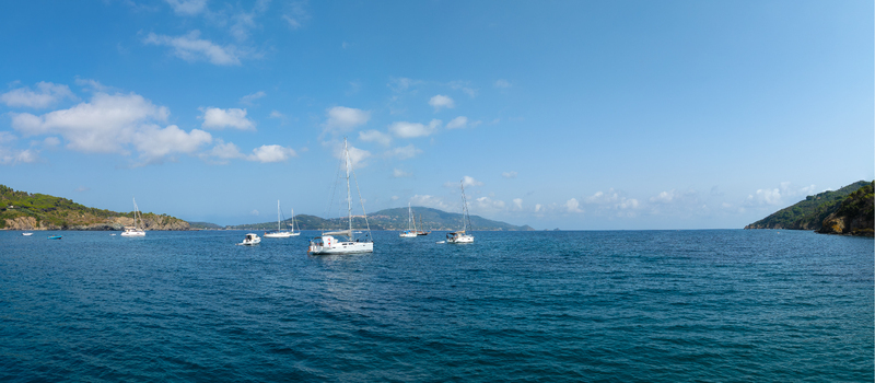 Aerial view of sailboats anchored in the calm blue waters of Lacona Beach surrounded by lush green hills under a clear sky in Lacona Tuscany Italy.