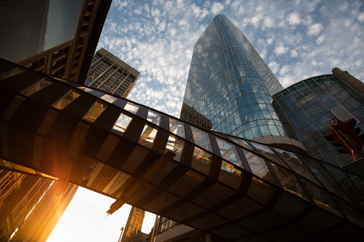 View of a modern glass skyscraper and a pedestrian skybridge under a blue sky with scattered clouds and bright sunlight flare in Toronto Ontario Canada.