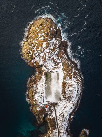 Aerial view of the snow dusted islet a stark contrast to the deep blue sea with a small red cabin nestled near a frozen pond Henningsv&aelig;r Nordland Norway.