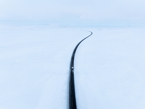 Aerial view of a lone car driving along a winding black asphalt road through a vast desolate snow covered landscape in Egilsstadir Mulathing Iceland. Aerial view of a lone car driving along a winding black asphalt road through a vast desolate snow covered landscape in Egilsstadir Mulathing Iceland.