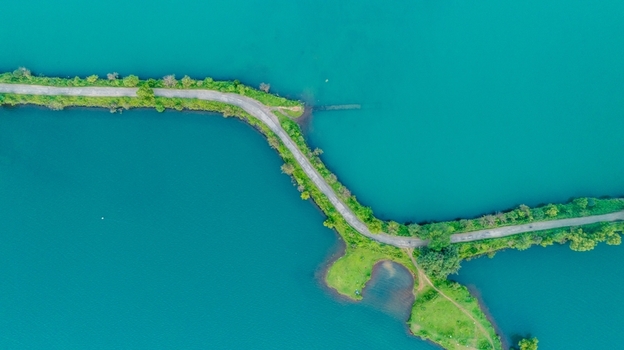 Aerial view of Upper Vaitarana Dam with a narrow road winding through vibrant turquoise reservoir waters surrounded by lush green vegetation in Ondli Maharashtra India.